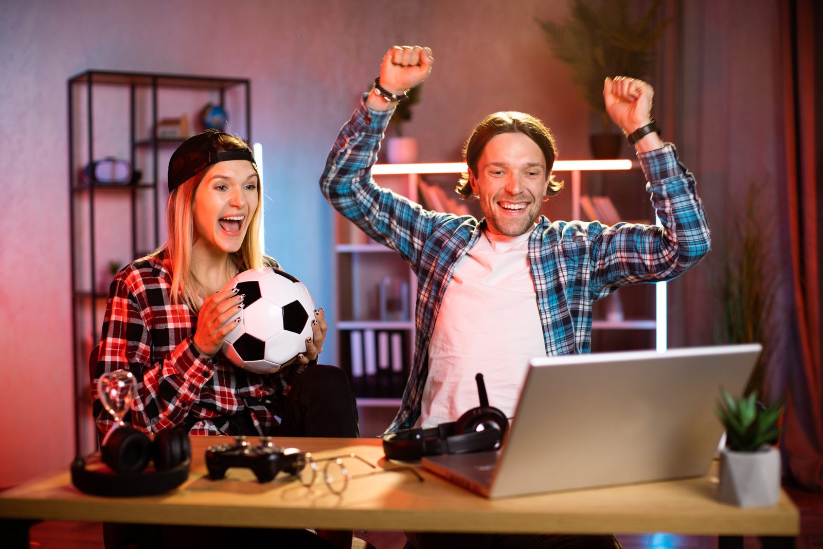 Positive young couple giving high five while sitting at table and watching football game on wireless laptop. Entertainment during evening time at home. Soccer fan concept.